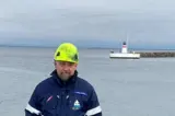 Man in blue jacket and green helmet in front of lighthouse