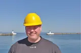 Man in gray shirt and yellow helmet in front of lighthouse