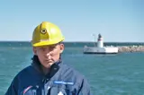 Man in blue jacket and yellow helmet in front of lighthouse