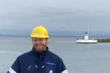 Man in blue jacket and yellow helmet in front of lighthouse